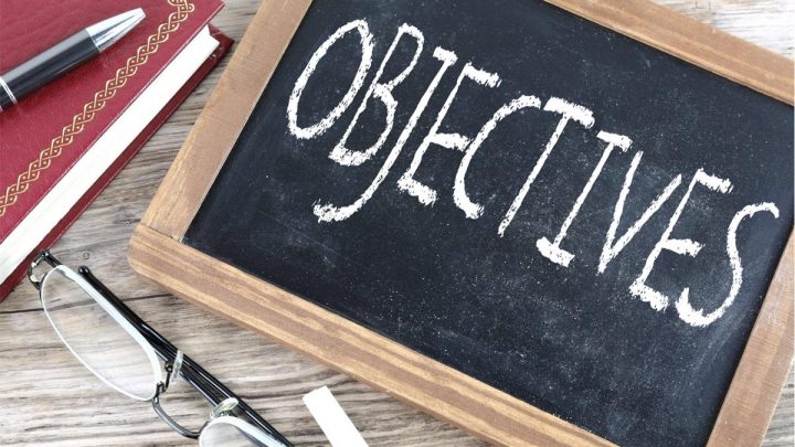 The word 'Objectives' written on a chalkboard, on a desk with a book, pen, and eye glasses.